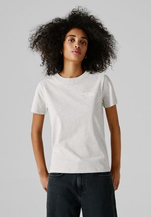 Žena s kudrnatými vlasy, oblečená v světle šedém tričku Red Bull Racing a tmavých džínách, stojící na pozadí jednobarevné světle šedé stěny.