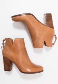 Stivaletti alla caviglia in pelle color cuoio con tacco spesso in legno, caratterizzati da lati traforati e dettaglio annodato sul retro. Texture liscia con finitura lucida.