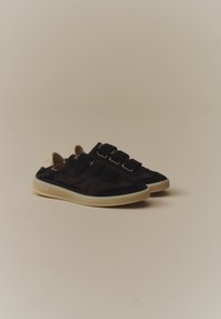 Paire de baskets basses noires avec trois bandes velcro et semelles en caoutchouc écru sur fond beige.