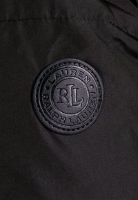 Svart tyg med en cirkulär, präglad läderpatch med texten "RALPH LAUREN" och "LAUREN", som detaljerar en texturerad kant runt omkring.