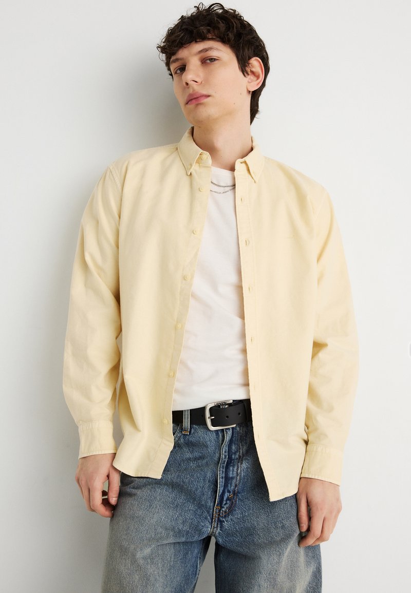 Jeune homme portant une chemise boutonnée jaune pâle par-dessus un t-shirt blanc, un jean bleu et une ceinture noire, debout devant un fond blanc uni.