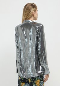 Silberne, metallische Langarmbluse mit glänzender Oberfläche, ausgestattet mit einem spitzen Kragen und Knopfmanschetten, die eine glatte, strukturierte Oberfläche zeigt.