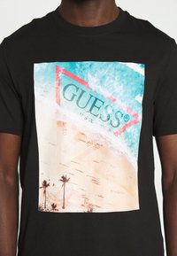 Camiseta de algodón negra con un gran gráfico temático de playa que presenta aguaturquesa, arena, palmeras y un logo triangular de "GUESS".