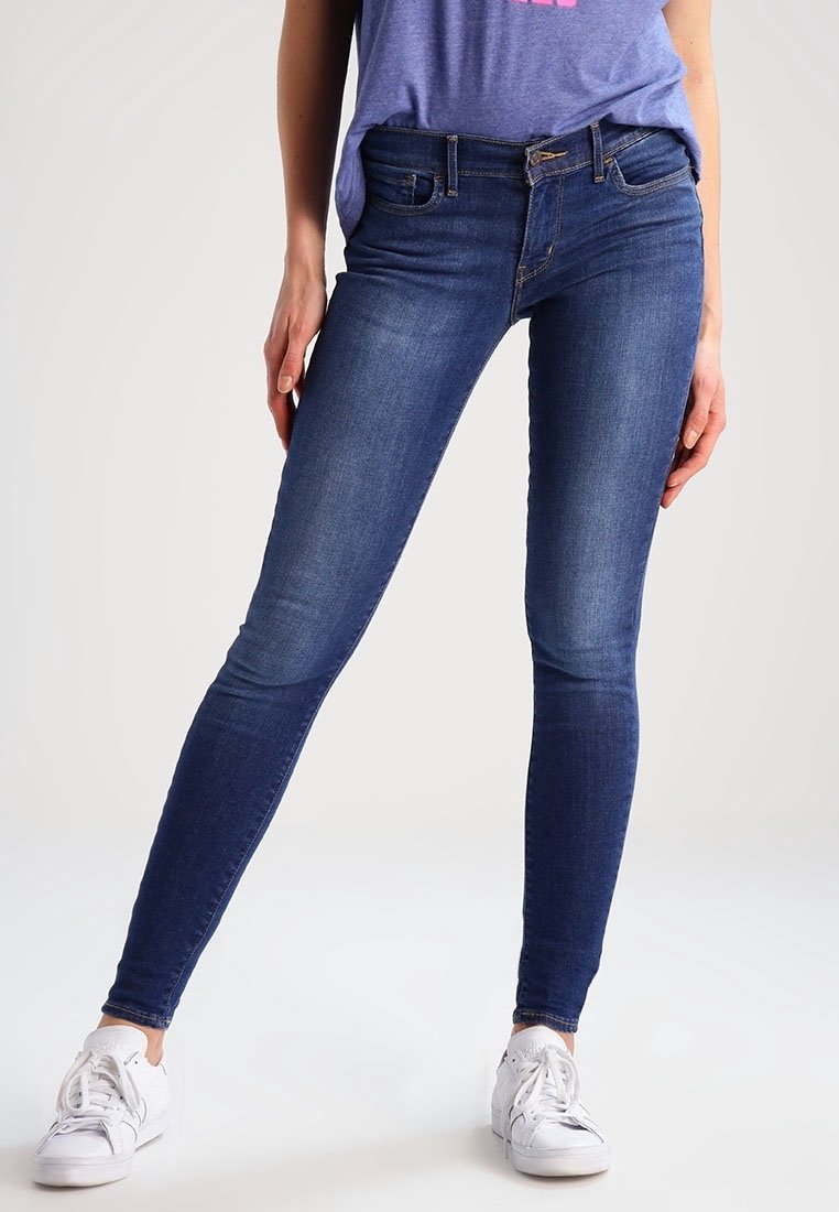 Femme portant un jean skinny bleu foncé, des baskets blanches et un t-shirt violet, debout, jambes légèrement écartées, sur un fond uni.