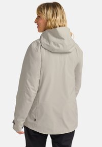 Femme aux cheveux blonds portant une veste à capuche gris clair et un pantalon noir, se tenant de dos avec la tête tournée vers la gauche sur un fond uni.