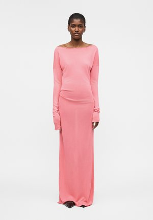 Γυναίκα που στέκεται φορώντας ένα μακρυμάνικο, μακρύ ροζ φόρεμα με λαιμόκοψη σκαφάκι και μαύρα μυτερά παπούτσια σε απλό λευκό φόντο.