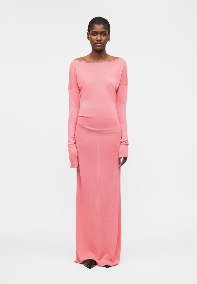 Γυναίκα που στέκεται φορώντας ένα μακρυμάνικο, μακρύ ροζ φόρεμα με λαιμόκοψη σκαφάκι και μαύρα μυτερά παπούτσια σε απλό λευκό φόντο.