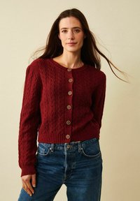 Körsbärsröd stickad cardigan med ett mönster av textur, rund halsringning och sju bruna knappar framtill. Ribbad fåll och ärmslut. Bärs tillsammans med blå jeans.