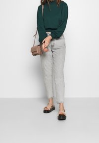 Grön långärmad topp med draperade manschetter, ljusgrå högmidjade jeans och tanfärgade sandaler med svart sula. Liten tanfärgad axelväska.