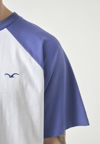 Short-sleeve raglan t-shirt featuring white body and purple sleeves, made of soft cotton, with a small purple logo on the chest.