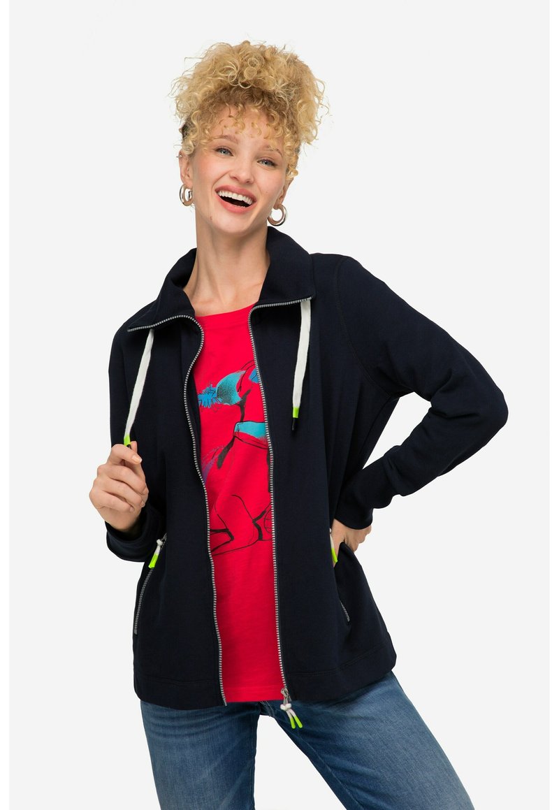 Chaqueta de mezclilla azul marino con cremallera en la parte delantera y cuello alto, con tiradores de cremallera verdes, combinada con una camiseta gráfica de color rojo brillante. Tela suave, corte relajado.