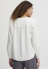 Camicia bianca in lino a maniche lunghe con orlo arrotondato, polsini con bottoni e dettaglio di cucitura sul retro. Il tessuto sembra leggero e texturizzato.