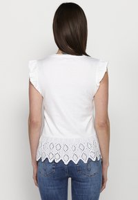T-shirt bianco in cotone con maniche corte a volant, con orlo in pizzo a occhio di pernice. Indossato con jeans di denim blu.