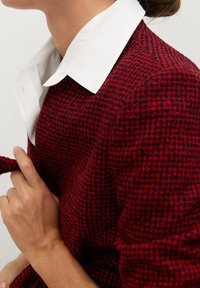 Sweter w wzór hounds-tooth w kolorze czerwonym i czarnym, z białą koszulą z kołnierzykiem. Posiada dopasowany krój i gładką teksturę.