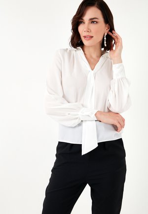 Weiße, transparente Bluse mit Ballonärmeln, einem Krawatten-Ausschnitt und geknöpften Manschetten, kombiniert mit schwarzen Hosen. Glatte Textur.
