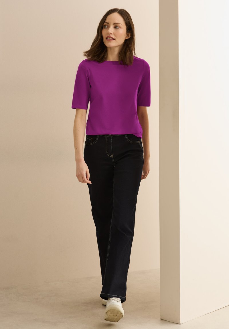 Fuchsia kortærmet bluse parret med mørkeblå jeans. Blusen har en rund halsudskæring; jeansene har synlige sømme og en afslappet pasform.