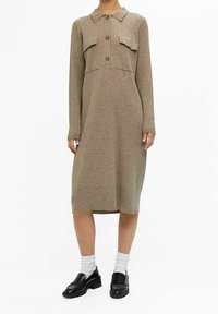 Robe beige en maille côtelée, manches longues, longueur au genou, avec col, trois boutons sur le devant, deux poches poitrine, portée avec des chaussettes blanches et des mocassins noirs.