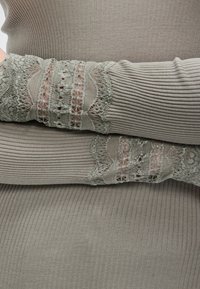 Άτομο που φοράει πουλόβερ πλεκτό ριμπ σε απόχρωση ταουπέ με λεπτομέρειες δαντέλας στα μανίκια, με τα χέρια σταυρωμένα πάνω στον κορμό σε φόντο από ύφασμα ταουπέ που ταιριάζει.