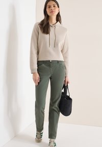 Femme portant un sweat à capuche beige, un pantalon vert olive et des baskets, tenant un petit sac à main noir sur un fond clair uni.