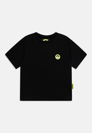 Camiseta negra de manga corta con pequeño gráfico de cara sonriente amarillo neón con colmillos en el pecho izquierdo y etiqueta neón a juego en el lateral.