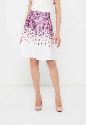 Femme portant une jupe blanche avec un motif de pétales floraux violets commençant à la taille, associée à des chaussures à talons hauts couleur nude.