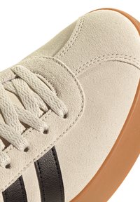 Närbild av en beige mockason sneaker med krämfärgade snörningar, svarta ränder och en brun gummisula synlig.