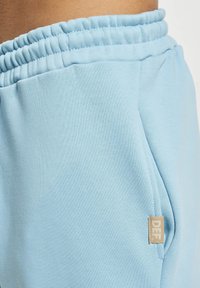 Pantaloni de trening de culoare albastru deschis, dintr-un material moale, cu talie ajustabilă prin șnur, buzunare laterale și un mic eticheta bej cu "DEF" pe lateral.