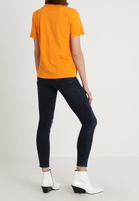 Camiseta de algodón naranja en una figura, combinada con jeans ajustados oscuros y botines blancos con tacón cuadrado. Diseño sencillo, colores sólidos.