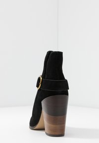 Botín de ante negro con puntera redondeada, detalle de hebilla lateral y tacón apilado estilo madera. Textura suave, diseño elegante.