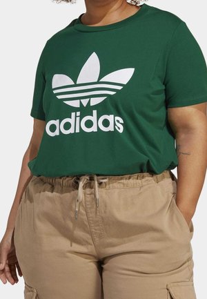 Person iført en grøn Adidas t-shirt med hvidt logo og beige snøre baggy bukser, hænder i lommerne, vist fra nakken til øvre lår.