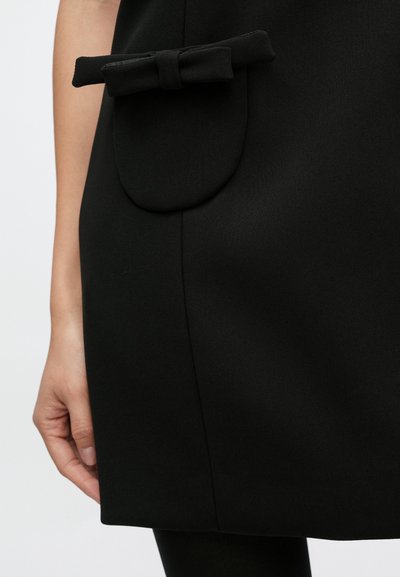 Fekete ruha, amelyen kör alakú zseb és masni díszítés található. Az anyag sima textúrájú. Egy kéz látható, amely tartja a ruhát.