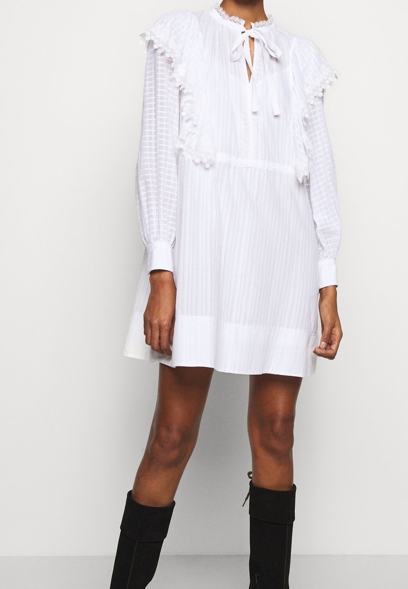 Hvid kjole med stribet tekstur, lange ærmer og flæseaccents. Har lukning med bindebånd i halsen og udvidet kant. Sorte knæhøje støvler.