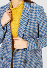 Modrá a béžová bunda s vzorem psí zub s černými knoflíky, nošená přes žlutý svetr s kabelovým vzorem. Vlna, klasický límec.