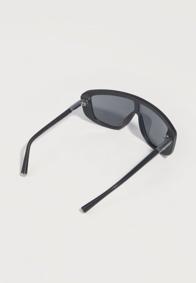 Μαύρα γυαλιά ηλίου τύπου wraparound με σκουρόχρωμους φακούς, φαίνονται από πίσω πάνω σε λευκή επιφάνεια.