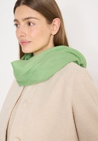 Femme aux cheveux châtain clair portant un manteau beige texturé et une écharpe vert clair enroulée autour du cou, regardant vers la gauche.