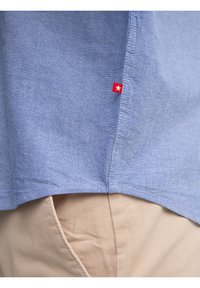 Prim-plan cu tivul unei cămăși albastru deschis cu o etichetă roșie care prezintă o stea albă, purtată peste buzunarul unei perechi de pantaloni bej.