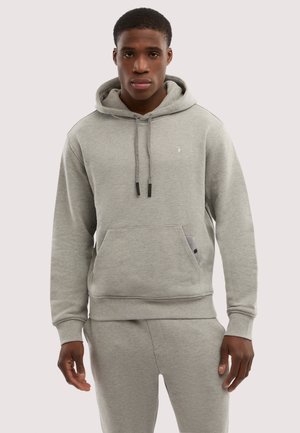 Ung mand iført lysegrå hættetrøje og matchende joggingbukser, stående mod en ensfarvet lyse baggrund.