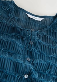Tyrkysová blůza s texturovanou třepenou tkaninou, má zapínání na knoflíky vpředu a skladaný design. Štítek říká "NEXT." Na materiálu je viditelný jemný lesk.