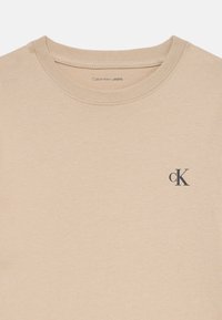Tricou din bumbac bej cu guler rotund, având un mic logo „ck” gri închis pe partea stângă. Textură netedă, design minimal.