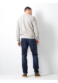 Vyras atsukęs nugarą, vilkintis šviesiai pilką džemperį, tamsiai mėlynus džinsus ir rudus batus prieš paprastą baltą foną.