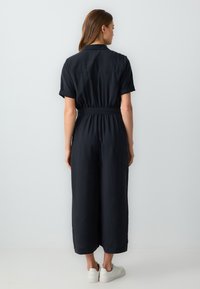 Combinaison noire à manches courtes, taille ceinturée, jambes larges et col. Le tissu semble léger et lisse. Portée avec des baskets blanches.