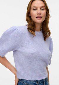 Lila pletena pulover s kratkimi pufastimi rokavi, okroglo ovratnikom in mehkim, strukturiranim zaključkom. Uporabljen z modrimi denim hlačami.