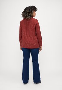 Blouse rouge à pois blancs, manches longues et pli au dos, associée à un jean bleu évasé et des chaussures noires.