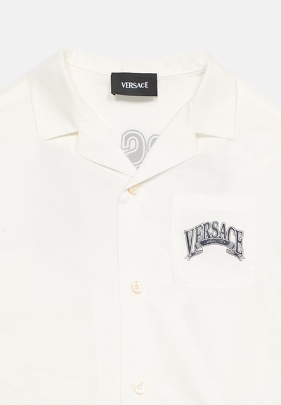 Λευκό πουκάμισο με κουμπιά και μαύρο λογότυπο "Versace" στην αριστερή τσέπη στο ύψος του στήθους και μαύρη ετικέτα μάρκας μέσα στον γιακά.
