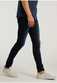 Tmavě modré džíny s úzkým střihem, hladkou texturou a minimem detailů, doplněné světlými teniskami.