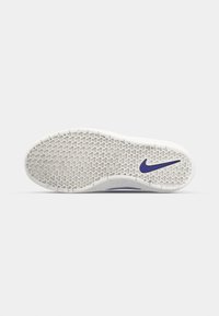 Semelle de chaussure blanche avec un motif hexagonal de crampons et un logo Nike Swoosh bleu foncé près du talon sur un fond uni.