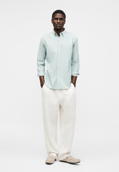 Mann står med hendene i lommene iført lys blå skjorte med knapper, løse hvite bukser og beige loafers mot en ensfarget bakgrunn.