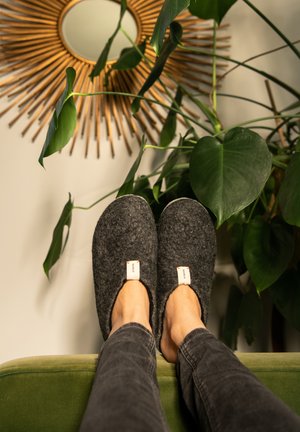 Šedé měkké pantofle s kulatými špičkami a bezobratým designem, s malými štítky na každé straně. Nohy spočívají na zelené pohovce, na pozadí jsou rostliny.