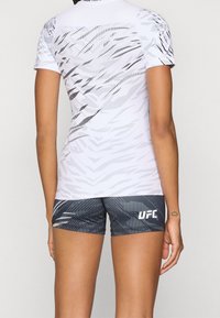 Bílé sportovní tričko s krátkým rukávem se šedým abstraktním vzorem a odpovídajícími šedými šortkami s logem "UFC". Materiál působí pružně a lehce.