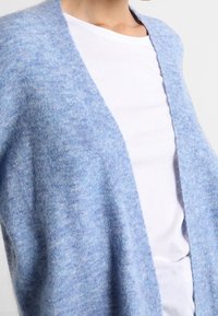 Osoba nosząca miękką, luźną, jasnoniebieską kardigan na prostym, białym t-shircie z okrągłym dekoltem na jednolitym tle.